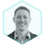 A man with short hair is smiling, wearing a checkered shirt and jacket. The grayscale photo, framed in a teal hexagon, reflects the confidence of AppSec Risk Prioritization for secure development.