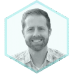 A smiling man with short hair and a beard, wearing a plaid shirt, shown in a black-and-white photo with a light blue hexagon border, represents expertise in Dynamic Application Security Testing (DAST).