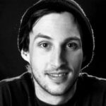 A young man with light skin, short dark hair, and slight stubble smiles gently. He wears a dark beanie and shirt against a plain black background. This black-and-white photo echoes the clarity of Shift-Left Security in CI/CD approaches.