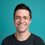 A man with short dark hair smiles, wearing a dark grey t-shirt, standing in front of a plain teal background—perfect for discussing Shift-Left Security in CI/CD or AppSec Risk Prioritization.