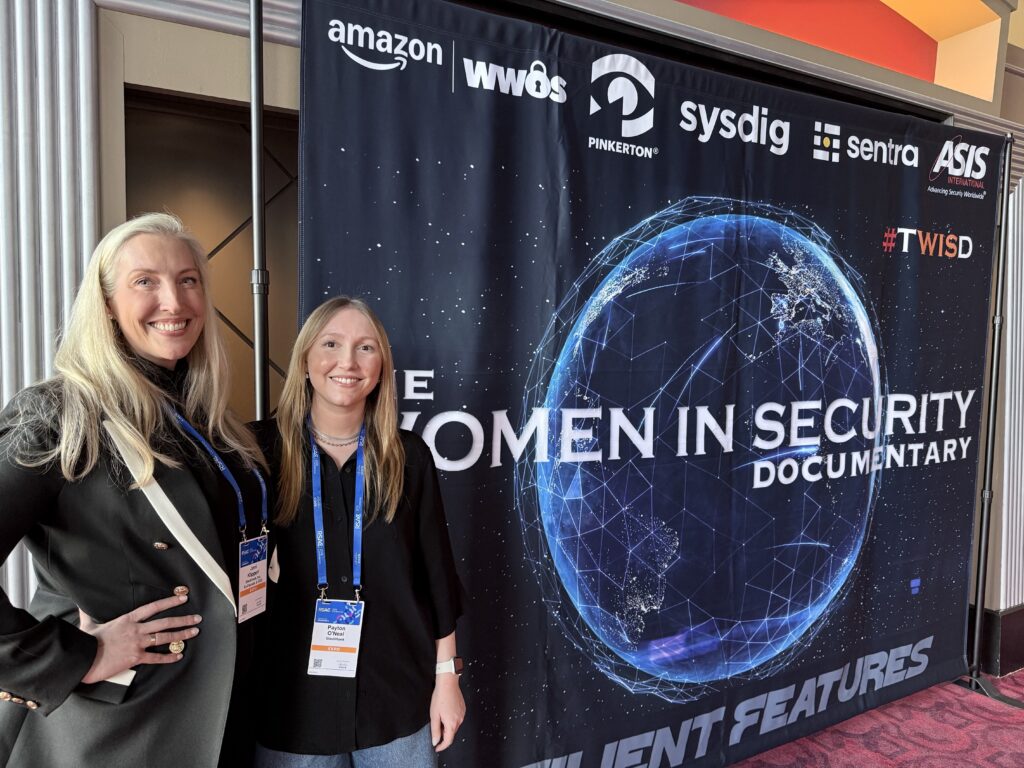 Joni Klippert and Payton O'Neal stand smiling in front of a banner that reads “Women in Security Documentary” with a digital globe and sponsor logos, including Amazon and Sysdig, displayed at the top.